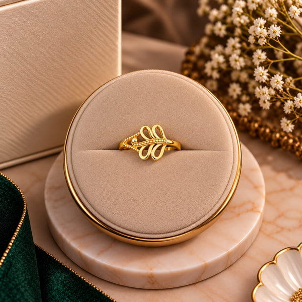 A gold ring with an elegant leaf-like design and small pavé accents, displayed on a round beige velvet ring box set on a marble coaster with decorative flowers and a green velvet ribbon.