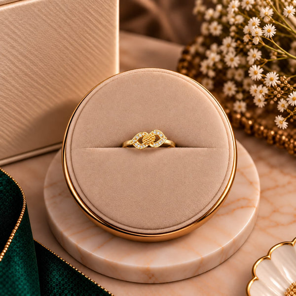 A gold ring with a knotted design and small clear stones, displayed in a round beige jewelry box on a marble-style stand with dried flowers in the background.