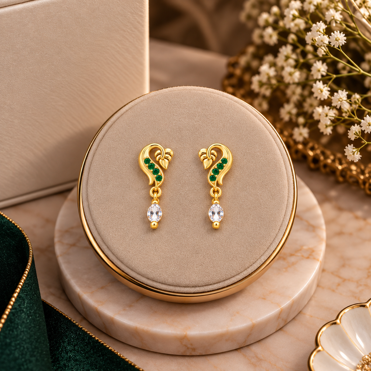 A pair of gold drop earrings with green stone accents and clear teardrop crystals, displayed on a round beige jewelry box lid over a marble stand with decorative flowers and ribbon in the background.