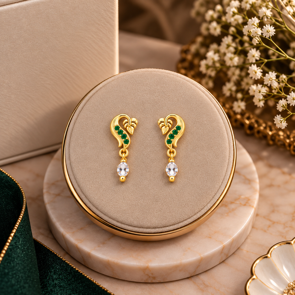 A pair of gold drop earrings with green stone accents and clear teardrop crystals, displayed on a round beige jewelry box lid over a marble stand with decorative flowers and ribbon in the background.