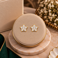 A pair of gold star-shaped stud earrings set with clear crystals, displayed on a round beige jewelry box lid with marble stands and floral decor in the background.