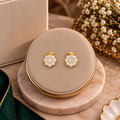 A pair of gold flower-shaped stud earrings with clear gemstones, displayed on a round beige jewelry stand with a marble base, styled with dried flowers and a green fabric corner.