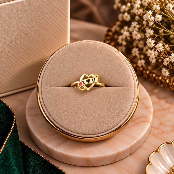 A gold ring with interlocking hearts and the word 'LOVE' in black enamel, accented with small red and green details, displayed on a round beige ring box.