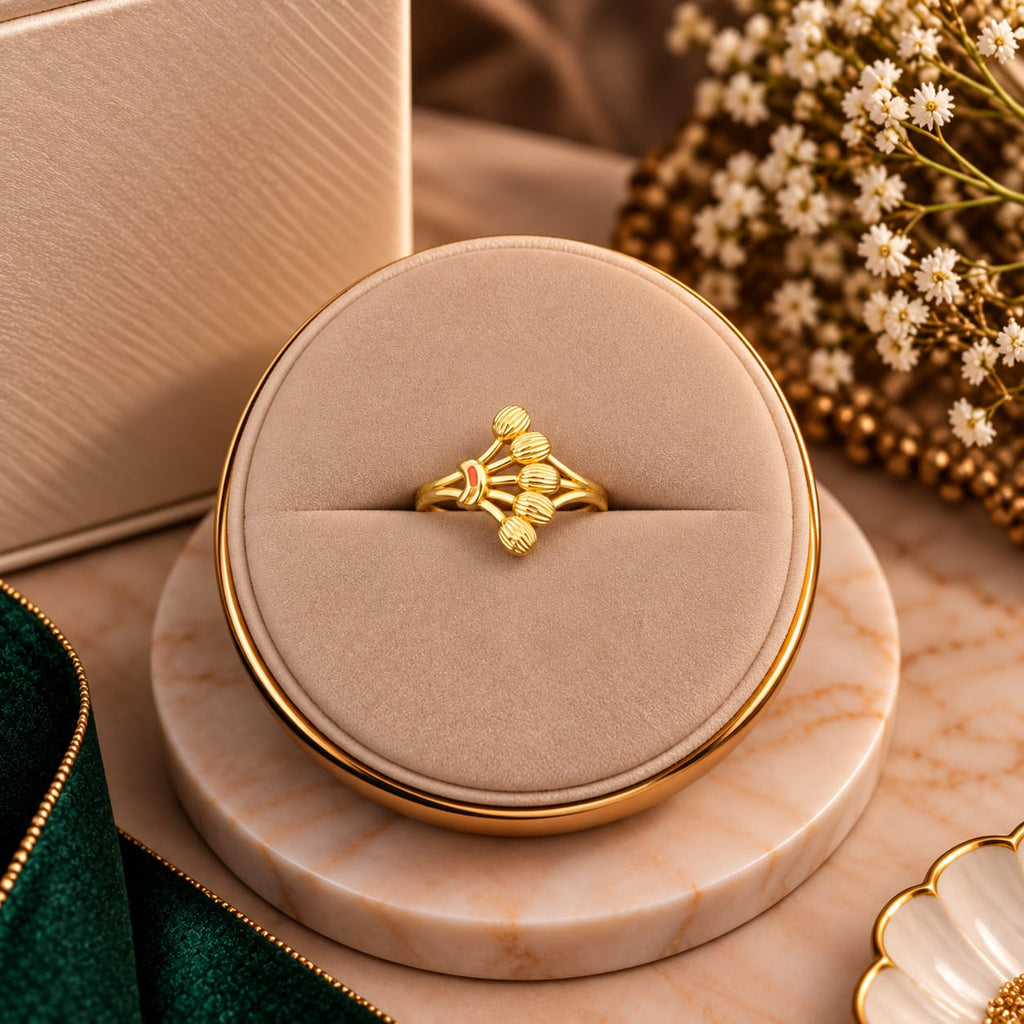 A gold ring with a textured floral or leaf-like design sits centered in a round beige jewelry display box on a marble-style base, with soft decorative props including dried flowers and a green fabric edge in the background.
