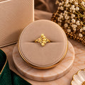 A gold ring with a textured floral or leaf-like design sits centered in a round beige jewelry display box on a marble-style base, with soft decorative props including dried flowers and a green fabric edge in the background.